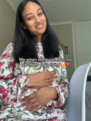 A woman with long black hair is smiling at the camera, with her hands gently resting on her pregnant belly. She is wearing a floral top. Text overlayed on the image reads "Me when someone asks me what I do for a living".