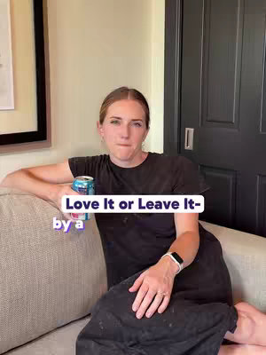 A woman sits on a beige couch, holding a can in her hand, wearing a black dress and a smartwatch. The background includes a framed picture and a dark door.