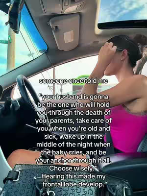 A woman is seated in the driver's seat of a car, seen from a slightly high angle. She is turned towards the passenger side, partially blocking her face with her hand. The background reveals a street with green street signs and buildings behind the car window. Text overlays appear on the image.