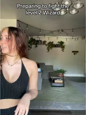 A young woman stands in the foreground, smiling towards the left. The background is a brightly lit room with decorative string lights and plants along a wall.