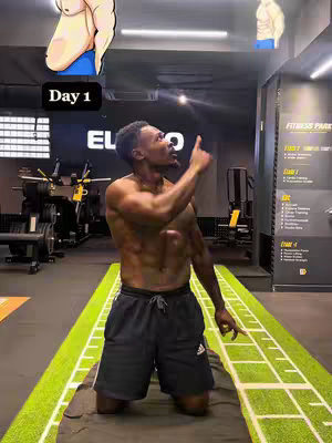 A man kneels on a green mat with an upright posture and a focused expression and points upwards with his right index finger. He is shirtless, showcasing his muscular physique and an Adidas logo on his shorts. The background features gym equipment and motivational signage.