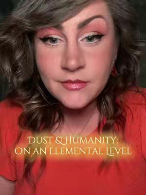 A close-up shot of a woman's face, looking at the camera with a gentle expression. The background is blurred, suggesting a shallow depth of field. Text overlayed at the bottom reads, 'DUST & HUMANITY: ON AN ELEMENTAL LEVEL.' The woman has styled, multi-toned wavy hair, with makeup, including pink eyeshadow and lipstick. Her top appears to be a bright orange color.
