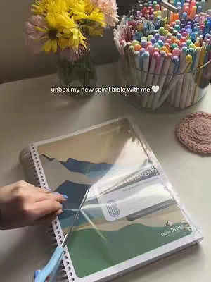 The cover image shows a scene with a table setting, featuring a spiral-bound Bible, a pair of scissors, a vase of flowers, and several containers of colorful pens. A hand is cutting the plastic wrapping off the Bible. The overall composition suggests organization and a workspace, possibly for journaling or study. The color palette is bright and inviting, with the focus on the Bible and the user's actions.