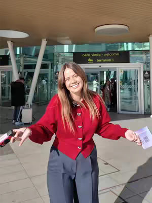 A woman stands smiling with arms outstretched in front of the Lisbon Airport terminal. She wears a red jacket and grey pants. The terminal entrance is in the background.