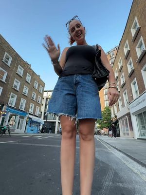 An eye-level shot of a woman standing on a street with buildings on either side. She is waving towards the camera. She is wearing a black top, denim shorts, and sunglasses, with a black bag on her shoulder.