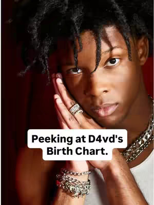 A close-up shot of a young Black man with short dreadlocks, looking directly at the camera with a slightly serious expression. He has a hand placed close to his face, with a ring on one finger and a bracelet on his wrist. A text overlay in a white rectangular box at the bottom of the image reads “Peeking at D4vd’s Birth Chart”. The background is a dark, blurred color of red.