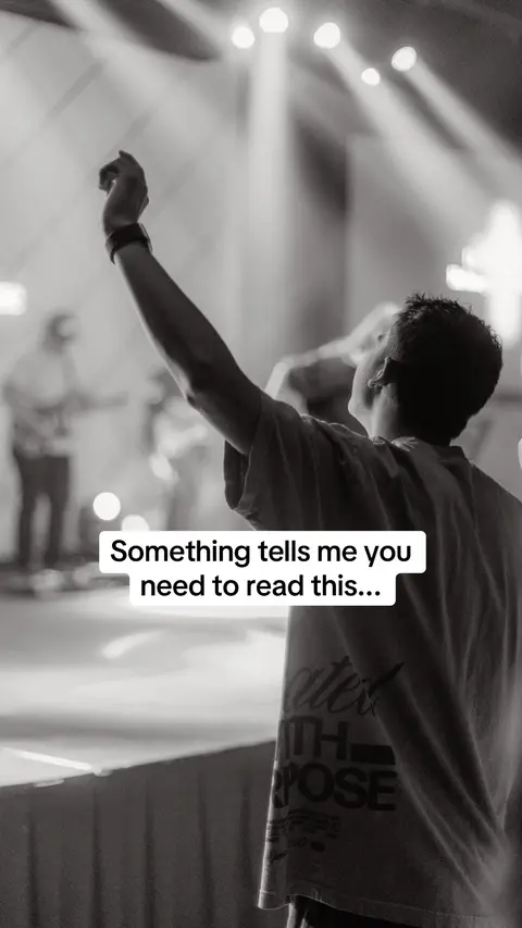 A black and white photo shows a person from the back with their right arm raised towards the upper right corner of the image. The person is in a crowd and the image has text overlay. The background is out of focus with stage lights visible.
