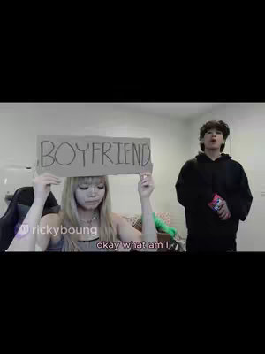 A woman holding a cardboard sign that says "BOYFRIEND" in front of a blurry background. Beside her stands another person in a black hoodie.