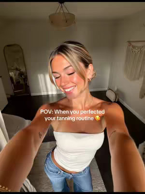 A selfie-style shot of a smiling woman indoors, appearing to be in a home. She is holding the camera at arm's length. The text "POV: When you perfected your tanning routine" partially obscures the image near her face.