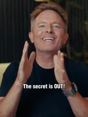 A man with light brown hair smiles broadly, his hands raised near his face as if applauding. He wears a black shirt and has a silver watch on his wrist.