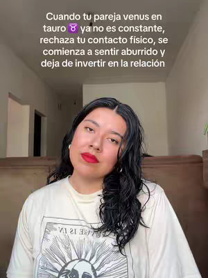 A woman with long dark hair and red lipstick is centered in a shot. She's wearing a printed t-shirt and leaning slightly, with text overlayed detailing astrological concepts. The background is softly lit with a wooden element visible.