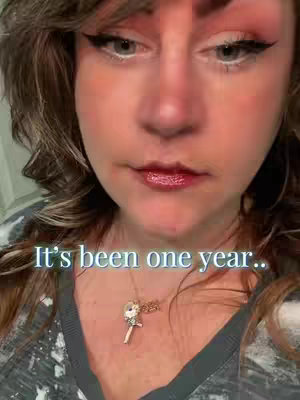 A close-up shot of a woman's face. Her face is centered in the frame. She has brown wavy hair, dark eyeliner, and a slight smile. There is a necklace with a charm and the text overlayed on top of the image reads: "It's been one year.."