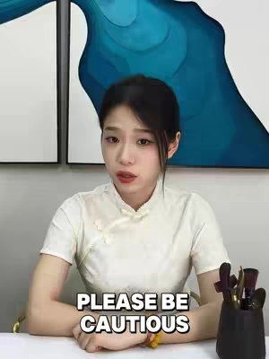 A woman with dark shoulder-length hair, wearing a white dress and speaking directly to the camera. A text overlay at the bottom reads "PLEASE BE CAUTIOUS". She has subtle makeup with red lipstick and light skin. The backdrop features abstract blue and white panel paintings, and on her right are makeup brushes in a holder.