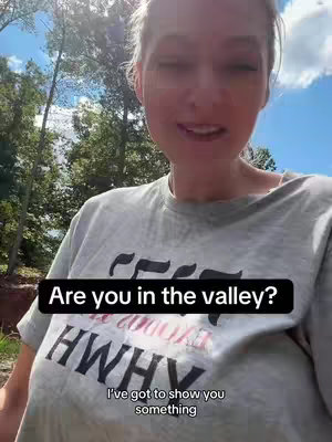 A woman in a grey t-shirt with black text stands and speaks towards the camera against a backdrop of trees and a partly cloudy sky.