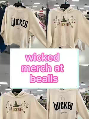 The cover image showcases four sweatshirts hanging on a rack. The sweatshirts are primarily off-white, each featuring a different design related to the musical 'Wicked'. A central, rectangular text overlay in pink displays the words 'wicked merch at bealls'.