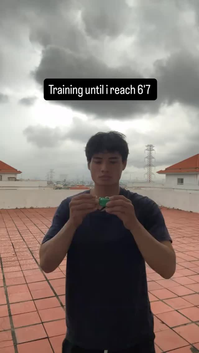 A young man stands on a rooftop, holding a small green object in his hands. He faces the camera with a neutral expression. The background consists of a cloudy sky and glimpses of buildings. A black text overlay reads 'Training until I reach 6'7'.