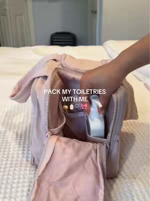 A light pink toiletry bag is partially unzipped, revealing the contents inside. A hand is reaching in to grab a white bottle. The background shows a bed with a white comforter and a room, and clothing.