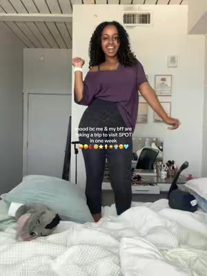 A young woman with curly dark hair is standing on a bed with white bedding. She's wearing a purple off-the-shoulder t-shirt and dark leggings. She has a bright smile, and her arms are raised in an excited gesture. A text overlay on the image states 'mood bc me & my bff are taking a trip to visit SPOT in one week' followed by a series of emojis.