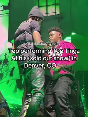 The image captures two individuals on a stage. The person on the left is wearing a black beanie, a black vest over a black shirt, and black pants, seen from the back. The person on the right of the frame wears a pink hoodie and black pants. Text overlayed indicates the performers are at a sold-out show in Denver.