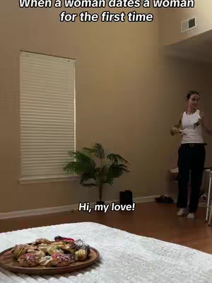 The cover image shows a domestic scene: a woman standing and smiling in a room with dark hardwood floors. In the foreground is a white comforter on which a plate of cookies is located. At the window, a blind is drawn, and light comes through it. The text overlay reads, "When a woman dates a woman for the first time" and "Hi, my love!".