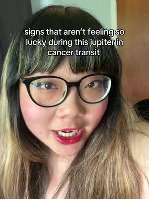 A selfie showing a young woman with fair skin, black-framed glasses, and long, highlighted hair . The woman has a slight smile and is looking directly at the camera. Text overlayed across the top of the frame reads: 'signs that aren't feeling so lucky during this jupiter in cancer transit'. The lighting is bright, and the background appears to be a nondescript interior.