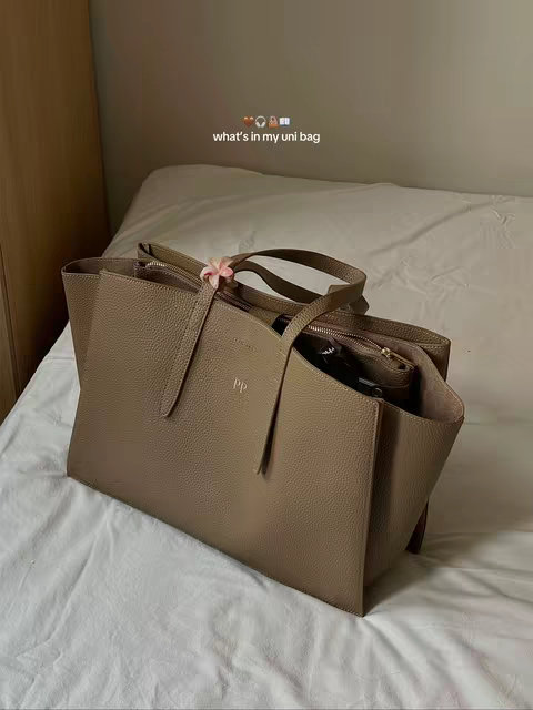 A large, neutral-toned tote bag sits open on a white bed. The focus is on the bag, highlighting its texture and shape.