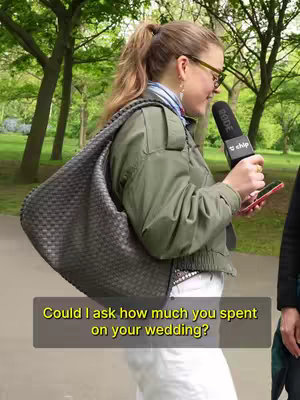 A woman in a green jacket and glasses stands outside, holding a microphone and a phone. She's positioned in front of a blurred green background with trees. A text overlay with the question "Could I ask how much you spent on your wedding?" placed at the bottom.
