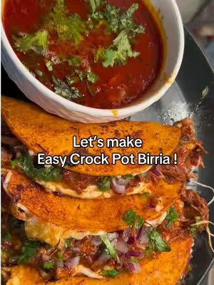 The image showcases a close-up of birria tacos and a bowl of consommé. Three tacos are stacked, filled with meat, cilantro, and onions, with a vibrant orange tortilla. A small white bowl holds a red consommé garnished with cilantro. The background is a black plate, with the composition highlighting the food's colors and textures.