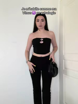 A woman stands centered, wearing a black tube top and pants, holding a black purse. The background is a plain white wall with a white door frame visible on the right. Text overlay in the upper left corner.