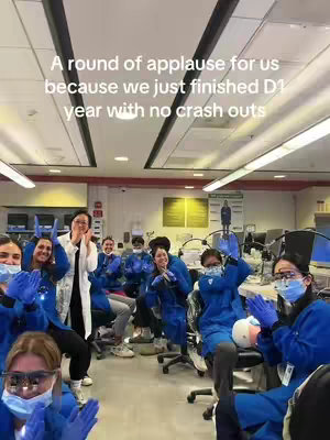 A group of dental students in blue scrubs are in a dental clinic setting. They are clapping enthusiastically toward the camera, with some wearing face masks and gloves. Text overlay at the top reads 'A round of applause for us because we just finished D1 year with no crash outs'.