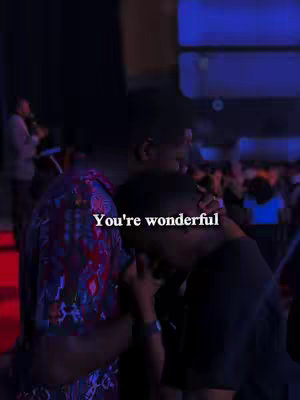 Two men are embracing in a dimly lit setting, likely an indoor venue, possibly a church or similar. The foreground features two men, one in a vibrant, patterned shirt and another in a black shirt, appearing to be hugging. One man is comforting the other by embracing and touching his shoulder. Soft lighting and blurred background elements such as other people or stage components give a calm mood. overlaid text that says: You're wonderful.