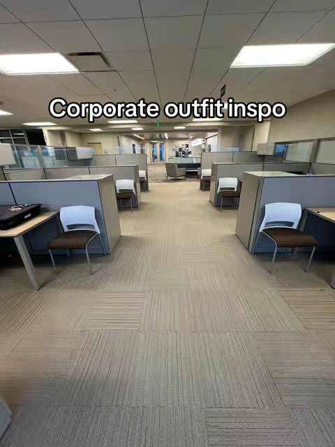 An office space with rows of cubicles and unoccupied desks. Cubicle walls are gray, and chairs at the desks have brown and white seats. Fluorescent lights are on the ceiling. The floor is covered with patterned carpet.