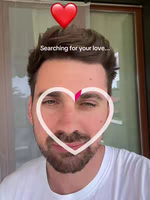 A selfie-style image of a man's face, framed from the chest up. The man has dark hair and a neatly trimmed beard. Overlayed on the image is a white heart outline around his face, a red heart icon above his head, and the text 'Searching for your love...' positioned between the two hearts. Behind the man is an outdoor scene, with a curtain and window visible.