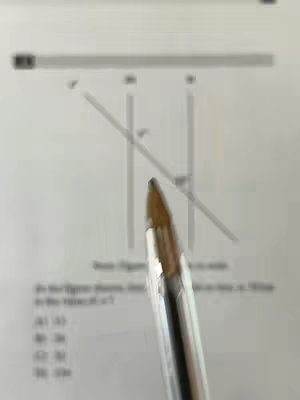 The image is a close-up of a pen, focused on its tip, pointing at a geometry problem written on a white paper, angled towards the center. Lines and some text including numbers are visible on the paper forming an outline of the figure. The background is blurred, which isolates the pen and the paper, making these elements in focus.