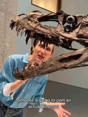 A woman with a surprised expression peers through the open jaws of a dinosaur skull. She is wearing a blue shirt and is positioned in front of a museum-like backdrop. Text overlayed at the bottom reads 'someone is going to own an actual Dinosaur'.