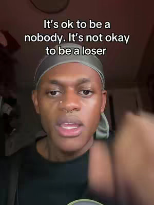 A person with dark skin and a nose ring looks directly at the camera, framed from the chest up. They wear a black shirt and a silver durag. Text overlayed on the top reads, 'It's ok to be a nobody. It's not okay to be a loser'. The background appears to be a room with a blurred, dark ceiling.