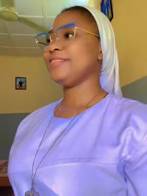 A woman wearing glasses, a scarf, and religious attire looks slightly off-camera in a medium shot. The background is a combination of yellow and blue walls. A framed art piece is visible on the yellow wall.