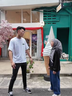 A man and a woman stand in front of a building with an ice-cream prop. The man in dressed in a simple white t-shirt and black pants. The woman wears a hijab and casual clothes. The setting appears to be an urban area with other decorative elements.