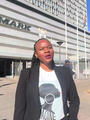 A woman with braided hair is standing and speaking in front of a large building with the words 'Department of Home Affairs, Head Office' visible. She is wearing a black blazer over a white t-shirt with a graphic on it. The composition is a medium shot.