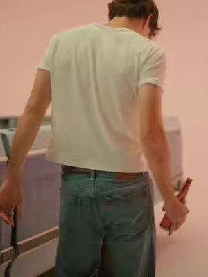 The image features a person from the back, wearing a white t-shirt and blue jeans, holding a bottle. They are standing in front of a pink backdrop with metal details on the lower left and a cooler on the right