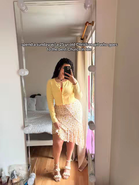 A woman takes a selfie in a mirror, standing in front of a bed. She wears a yellow sweater, a patterned skirt, and sandals. The mirror has lights around it. The background shows a bed and other decor.