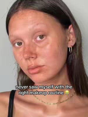 The cover image is a close-up of a young woman's face. She has visible skin texture and some freckles with dark hair partially obscuring her forehead. She is wearing a black top and a thin gold necklace with a small pendant. Her expression is slightly subdued. The background is a plain, light color, possibly a wall. The text overlay near the image bottom reads: "never saw myself with the right makeup routine 🥺"