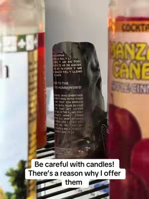 The image features a close-up of three glass bottles. The foreground includes a mostly obscured, cylindrical dark bottle holding what appears to be a partially melted candle. Behind it, on the right, is a bottle with red liquid and text, 'Manzana Cane', above the candle. On the left, another bottle, also with red liquid, supports the candle. The bottles are set on what appears to be a black and silver striped surface. Below the bottles, there's a white text overlay: 'Be careful with candles! There's a reason why I offer them'.