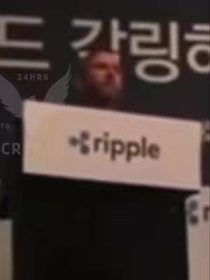 A person is seen in the background, behind what appears to be a sign that reads "ripple" in black letters with a symbol next to it. The sign is held up in front of the person and is white. Parts of black objects exist in the foreground and the text and background appear to be in a blurred.