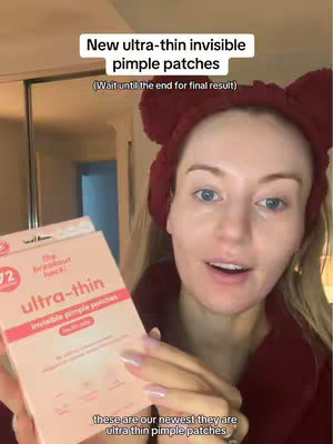 A woman with fair skin and blonde hair is centered in the frame, wearing a maroon robe and a headband with bear ears. She is holding a pink and white box of "ultra-thin invisible pimple patches". The background is a neutral room with a closet.