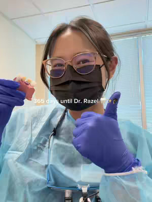 A selfie-style image featuring a woman wearing glasses, a black face mask, and blue surgical gloves. She holds a dental mold in her left hand and gives a thumbs-up with her right hand. The background is a dental office setting, with overhead lights and partial blinds visible.