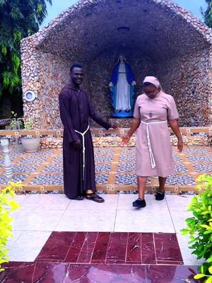 The cover image shows a religious scene with two people in front of a structure featuring a statue. A fair-skinned man dressed in a dark brown robe with a white cord stands to the left facing forward and smiling slightly. A woman dressed in a pink outfit with a white veil and belt is to the right, facing the man. Behind the people is an alcove containing a statue of the Virgin Mary with a blue robe.