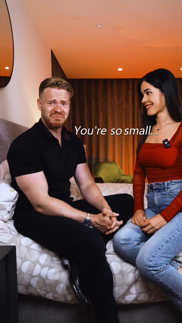 A cover image featuring two individuals seated on a bed. On the left, a man with short, reddish-blonde hair, wearing a black polo shirt, looks distressed. On the right, a woman with long black hair, wearing a red top and jeans, smiles at the man.