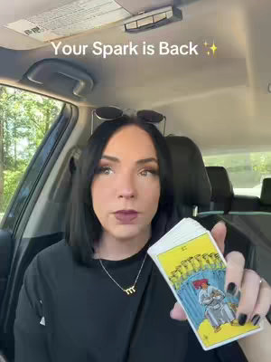 A woman sits inside a car, holding a deck of tarot cards. She is looking at the camera and has text on the image that reads: 'Your Spark is Back'. The background shows an interior car setup and a glimpse of the outside environment through the car window.
