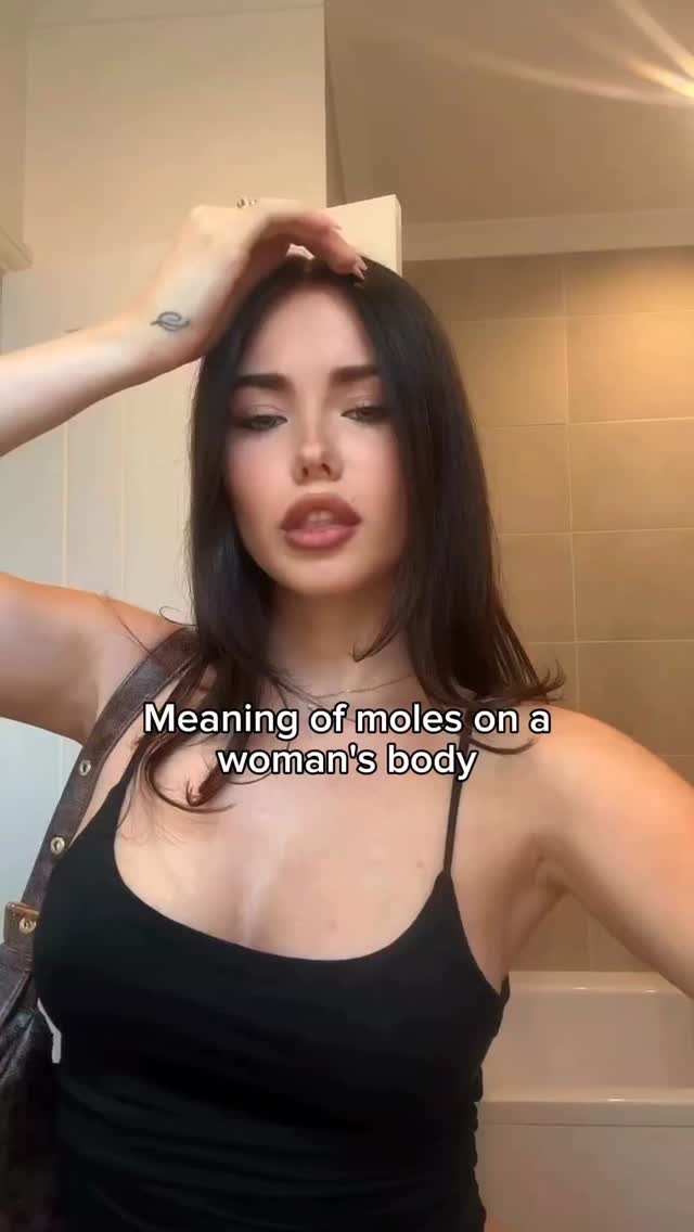 The cover image shows a young woman with long dark hair, wearing a black tank top. The woman is looking at the camera with a neutral expression, and has one hand in her hair. The text overlay reads 'Meaning of moles on a woman's body'. The background appears to be inside a bathroom.
